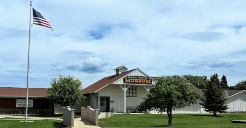 Exterior of the Central Montana Museum with an American flag, surrounded by trees and a cloudy sky.